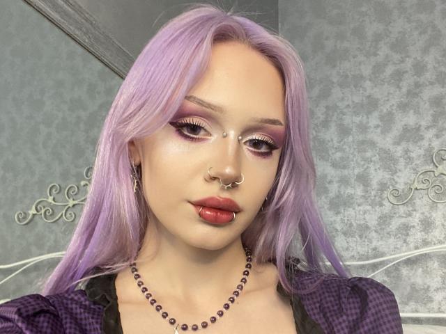 LiliClem has lavender hair, pink eyeshadow, a nose ring, and a lip ring with her tongue out. She's wearing a purple plaid shirt and a bead necklace.
