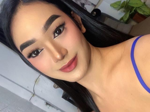 AsianHugeCockxX with dark eyebrows, long eyelashes, pink blush, red lipstick, and blue spaghetti strap, smiling at the camera, showing a cute face and straight black hair.