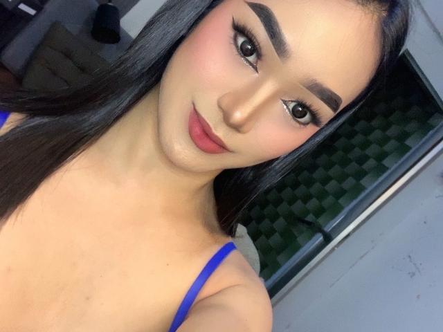 AsianHugeCockxX with long black hair, thick eyebrows, eyeliner, pink blush, red lipstick, and blue top, smiling at the camera, showing a cute face and small tits.