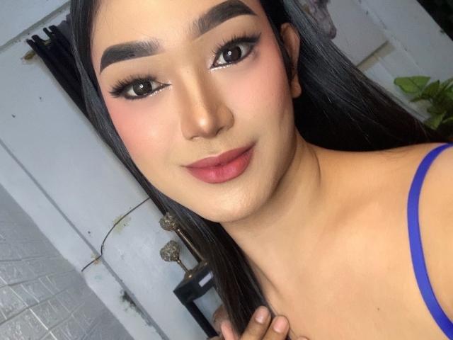 AsianHugeCockxX with dark eyebrows, black eyeliner, pink blush, red lipstick, and blue top, smiling at the camera, holding her collarbone. Her skin is smooth, and she has long black hair.