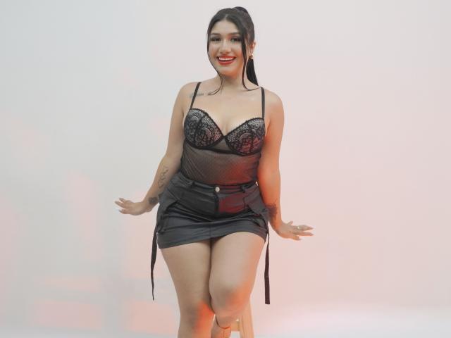 CamilaVerone has dark hair, red lipstick, and wears a black lace bra and sheer top with a black leather skirt, sitting with hands on knees, showing her tits and ass.