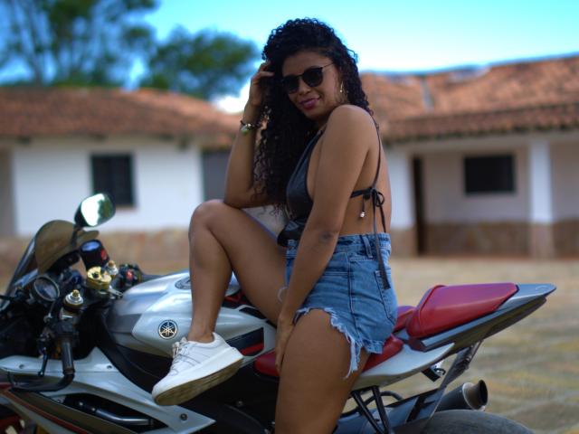 AnelicaSexos with curly black hair, wearing dark sunglasses, a gray bikini top, and frayed denim shorts, sitting on a red-and-silver motorcycle, showing her tanned thighs and white sneakers.
