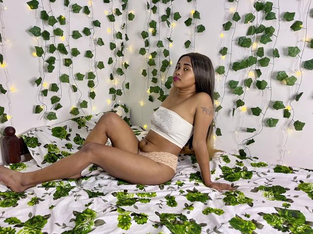 LaCoquineX with brown skin, long dark hair, small tits in white strapless top, pink lace panties, sitting on green leaf-patterned bed, surrounded by fairy lights and leaf garland.