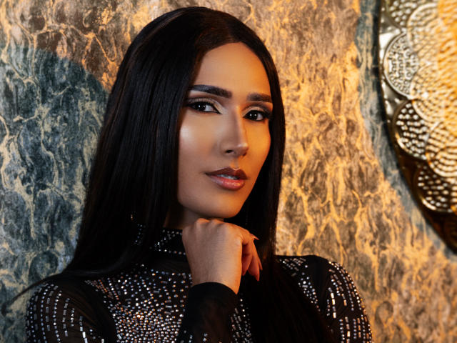 FallonRogers with long black hair, dark eyes, and shiny lipstick, wearing a black, sparkly top, touches her chin, looking sexy with a confident, sultry expression.