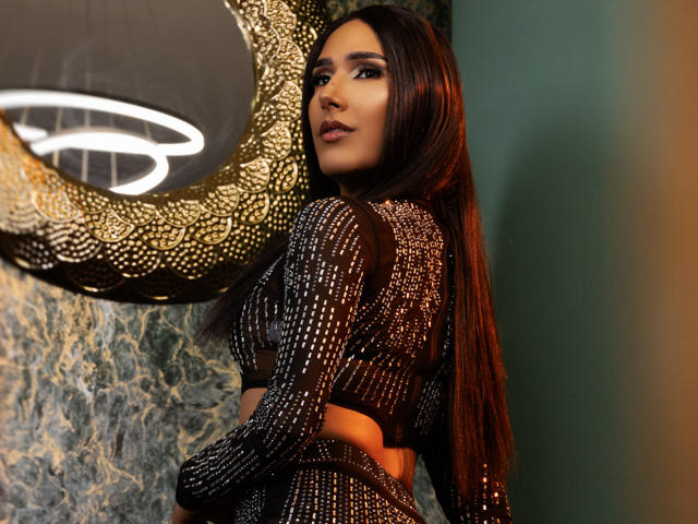 FallonRogers with long brown hair, wearing a black, sparkly, long-sleeve crop top and matching skirt, standing against a textured wall, looking over her shoulder.