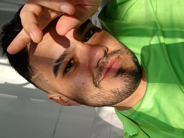 StevenHills with a green shirt, short black hair, and a beard, holding his head with sunlight casting shadows on his face, looking slightly to the side, smiling subtly.