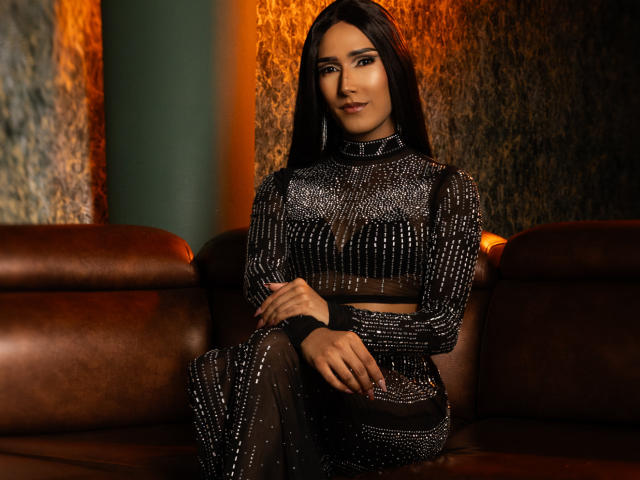 FallonRogers, with long dark hair, sitting on a brown leather sofa, wearing a black, sheer, sparkly, long-sleeve top and matching skirt, showing her tits and ass subtly.