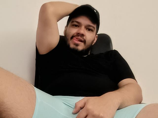StevenHills, a bearded man with a black cap, black shirt, and light blue shorts, is sitting with one arm behind his head, showing his relaxed, confident smile.