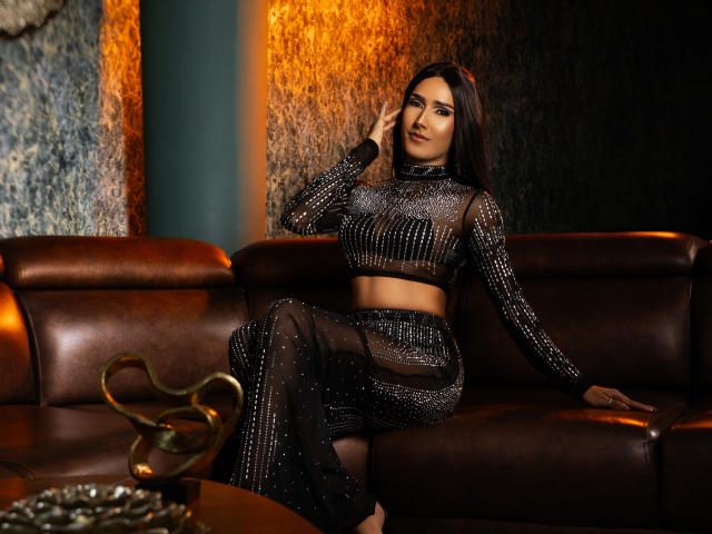 FallonRogers, dark-haired, wearing a black, sheer, sparkly crop top and skirt, sitting on a brown leather couch, hand near face, looking confident, with a gold decorative item in front.