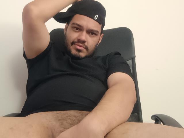 StevenHills, a chubby man with a black cap and beard, is sitting in a chair, legs spread, showing his hairy, flaccid, uncircumcised penis. He's wearing a black shirt.
