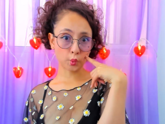 Emma2202 with curly brown hair, glasses, and pink pout, wearing a sheer black top with white daisy patterns, touching her cheek, surrounded by red heart-shaped lights.