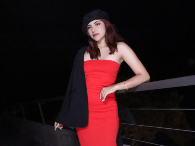 SpencerLe, vêtue d'une robe rouge sans bretelles, d'un béret noir et d'une veste noire, debout, une main sur la hanche, sur fond de nuit sombre, portant un bracelet, regardant la caméra..