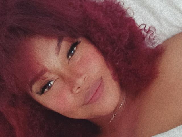 DalilaHeinz has curly red hair, dark eyebrows, black eyeliner, and a subtle smile with full lips. She's wearing a thin silver necklace, and her skin is freckled.