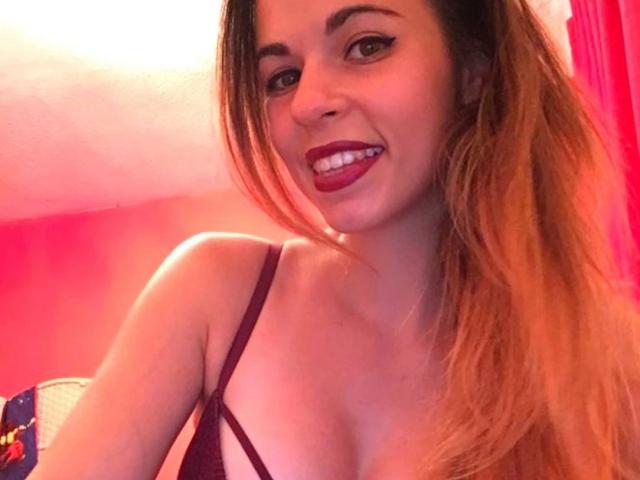 Nekane, con el pelo largo y castaño, lápiz labial rojo, sonriendo, vistiendo un sujetador morado, mostrando sus pechos en una habitación rosa..