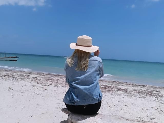 HaileyDavies with blonde hair, wearing a beige hat and blue denim jacket, sitting on a log, facing a turquoise ocean, her black ass visible.