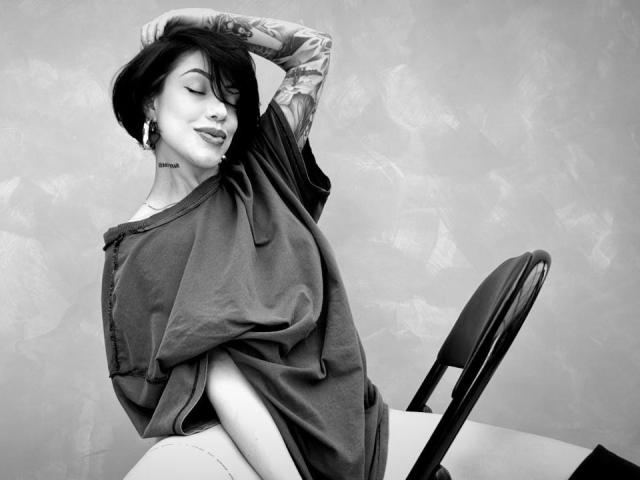 Black-and-white photo of HannaBorbon with tattoos, sitting on chair, wearing loose shirt, hand in hair, eyes closed, slightly smiling, showcasing small ass and visible sideboob.