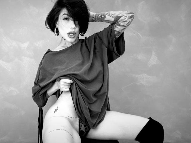 HannaBorbon with short dark hair, tattoos on arms, large black ear gauges, wearing oversized shirt, lifting shirt to expose small tattoo on left hip, sitting with legs apart.