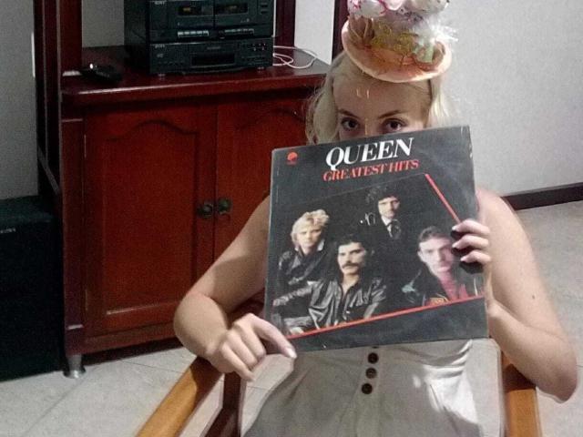 HaileyDavies with blonde hair, wearing a hat and white dress, holding a Queen "A Queen Is Born" CD, covering her tits. Wood cabinet and electronics in the background.