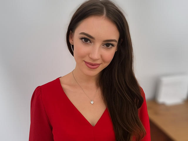 HarperSensitive has long brown hair, wearing a red V-neck dress, smiling with makeup, small silver necklace with a heart, and is standing with a subtle, sexy look.