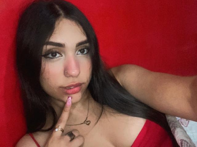 EmmaRubis with long black hair, nose ring, and pink lipstick, touching her pink lips with her finger, wearing a red top, against a red background.