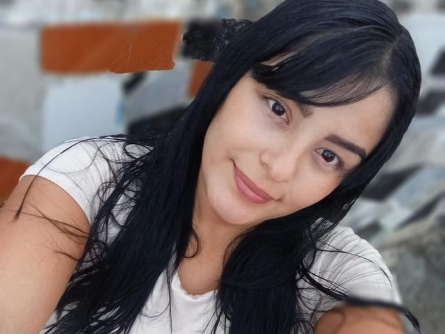 TalianaSquirt with black hair, brown eyes, and a white shirt, smiling softly at the camera. Her medium-sized, natural tits are subtly visible. She has a cute, relaxed look.
