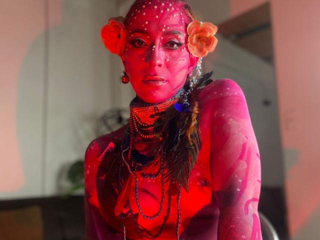 Sahrataylorr has red-pink body paint, orange flower earrings, and beaded necklaces, with sparkles on face and chest, looking at camera with serious expression.