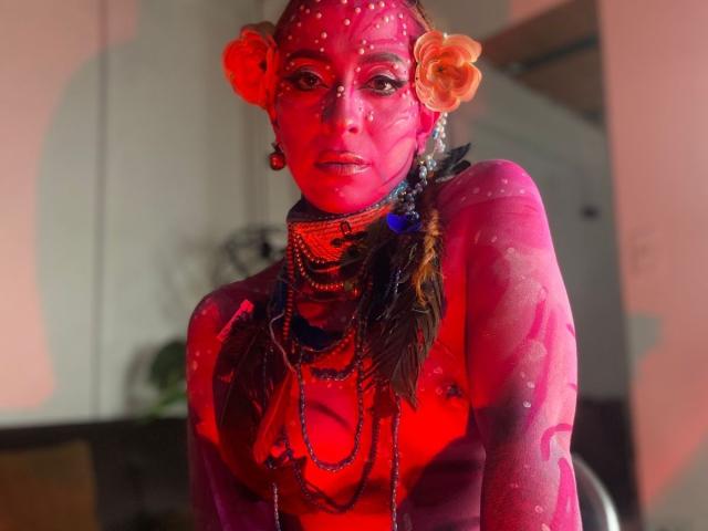 Sahrataylorr with bright pink body paint, orange flower in hair, and multiple bead necklaces, looking at camera. Painted face with white dots, pink skin, and dark feathers.