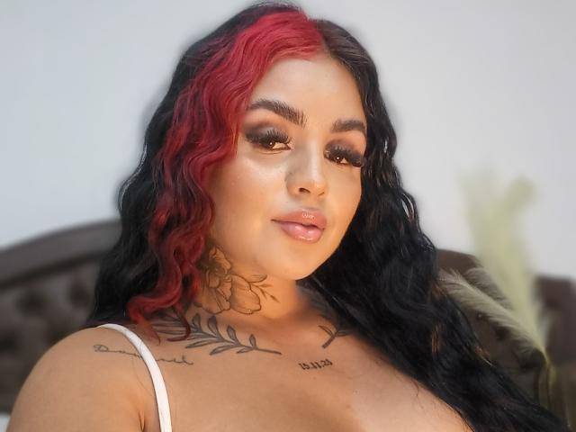 Gabrielle King, con capelli neri e rossi ondulati, sopracciglia folte, labbra lucide, tatuaggi sul collo e sul petto, indossa un reggiseno bianco, guarda la fotocamera, luce soffusa, con un leggero sorriso..