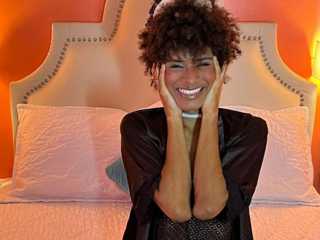 Violet Salvatore, con cabello castaño rizado, sonriendo, con las manos en las mejillas, vestida con una bata de satén negra, un cojín rosa y una pared naranja de fondo, sentada en una cama..