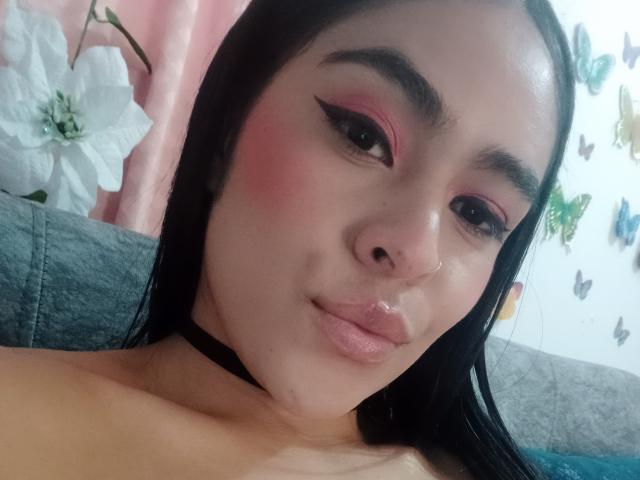 StellaBeckker tiene sombra de ojos rosa, delineador de ojos negro y un collar negro. Su cabello negro está suelto, está haciendo pucheros con un piercing en la nariz y tiene las mejillas rosadas. Sus senos no son visibles..