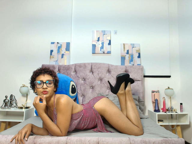 GraceTurnner with curly brown hair, glasses, and red lipstick, laying on a bed in a sparkly pink dress, black high heels, and her ass up, resting on her elbows.
