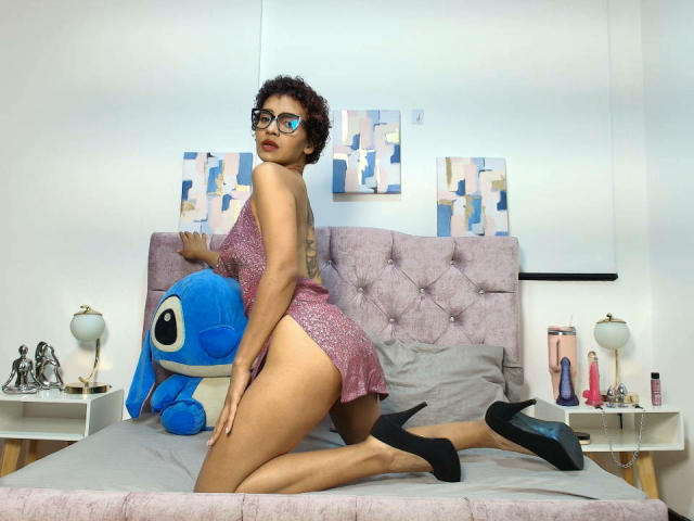 GraceTurnner with curly brown hair, glasses, pink sparkly dress, black heels, kneeling on bed, blue Stitch toy, exposing her ass, tattoo on back, white bedroom, modern art.
