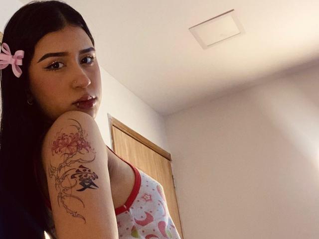 AnnaTorress, con cabello oscuro, un lazo rosa, un piercing en la nariz y un tatuaje en el brazo, vestida con una blusa colorida y estampada, mirando por encima del hombro, en una habitación con luz tenue..