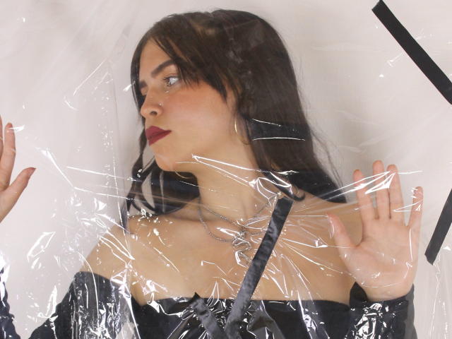 AgathaaQueen, con i capelli scuri, il rossetto rosso e l'abito nero con le spalle scoperte, è intrappolata in una plastica trasparente, con le mani alzate, che guarda di lato, mostrando il seno e un viso carino..
