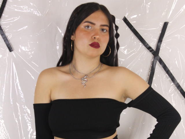 AgathaaQueen, com cabelo escuro, batom vermelho, blusa preta com um ombro descoberto, brincos de argola dourados e colar de prata, em pé contra um fundo branco de plástico. Seios visíveis, mão na cintura..