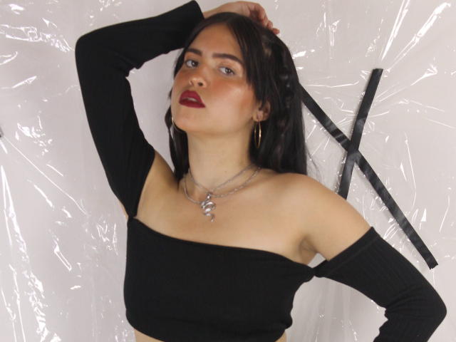 AgathaaQueen tem cabelo escuro, batom vermelho e uma blusa preta com um ombro descoberto. Ela está usando um colar e brincos de argola dourados, com um braço levantado. Os seios estão visíveis..