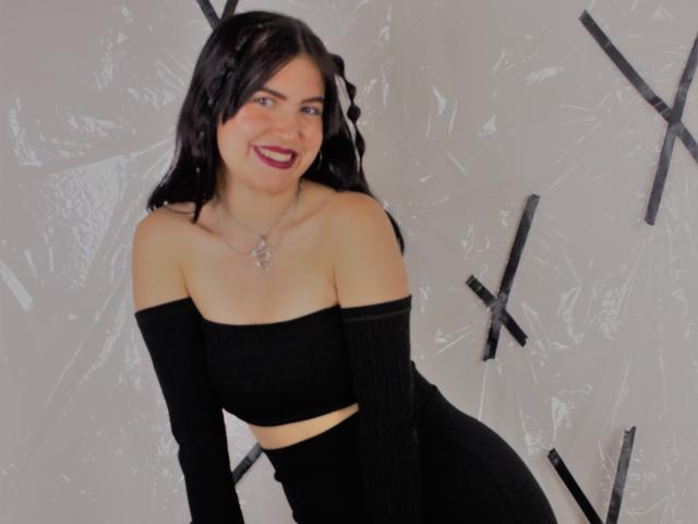 AgathaaQueen, con cabello negro, sonriendo, vestida con una blusa y pantalones negros de corte asimétrico, mostrando sus pechos y glúteos, con un fondo cubierto de plástico..
