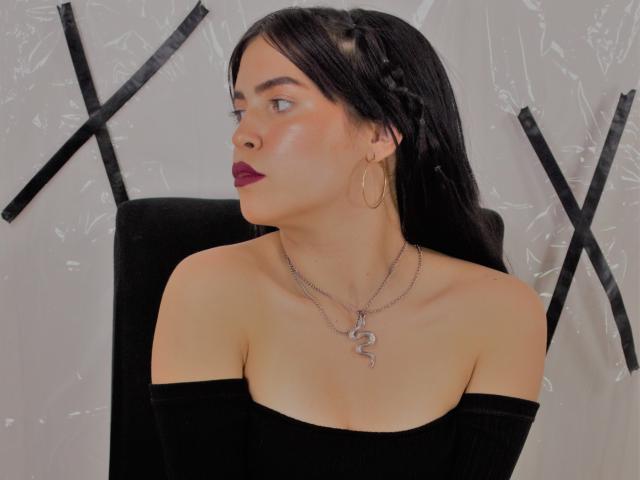 AgathaaQueen con i capelli scuri, intrecciati, indossa una maglietta nera a spalle scoperte, rossetto rosso, orecchini a cerchio dorati, una collana d'argento a forma di serpente, guarda di lato, pelle chiara, leggero trucco sulle guance, illuminazione soffusa..