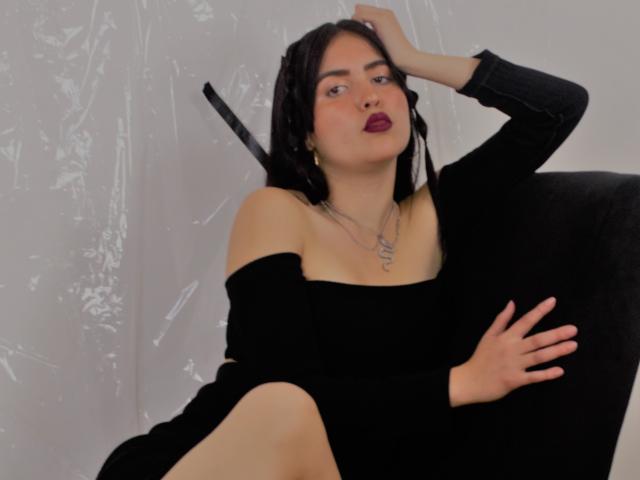 AgathaaQueen com cabelo escuro, vestido preto com um ombro descoberto, sentada em uma cadeira preta, com a mão na cabeça, perna levantada, olhando para a câmera, batom vermelho, colar, confiante, sexy..