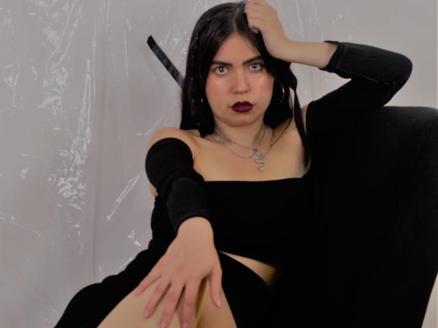 AgathaaQueen con i capelli scuri, un abito nero a spalle scoperte, rossetto viola e orecchini a cerchio, una mano sulla testa, gambe divaricate, che mostra la sua vulva, su uno sfondo ricoperto di plastica..