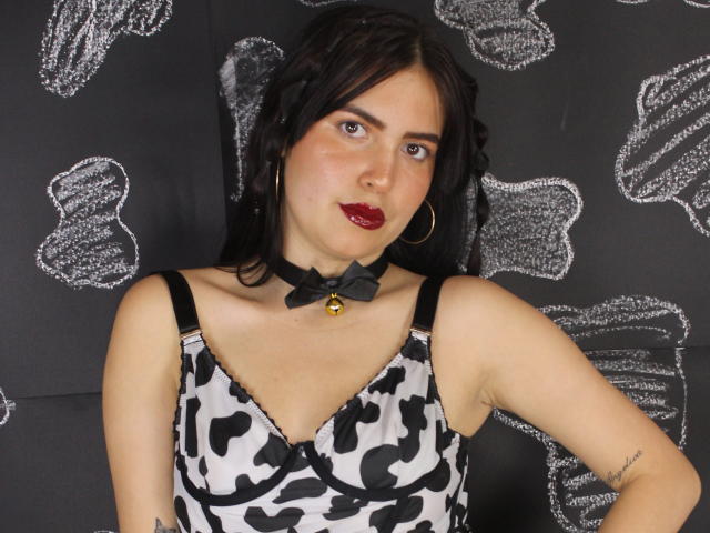 AgathaaQueen, com cabelo escuro, batom vermelho e um vestido com estampa de vaca em preto e branco, usando um colar preto com um sino e brincos de argola dourados, olhando para a câmera..