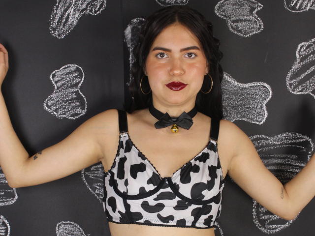 AgathaaQueen mit dunklem Haar, rotem Lippenstift und Reifenohrringen, trägt einen schwarzen und weißen BH mit Kuhmuster, eine schwarze Choker-Kette mit Glöckchen, die Arme sind erhoben, wobei kleine Brüste und Tattoos zu sehen sind..
