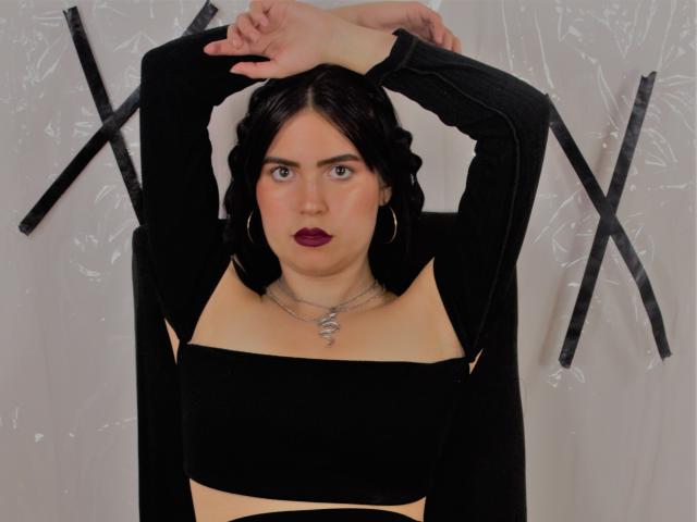 AgathaaQueen, con cabello oscuro, lápiz labial púrpura, top negro con aberturas, brazos levantados, mostrando sus senos, vistiendo un collar de plata y aros grandes, sobre un fondo blanco con letras "X" negras..