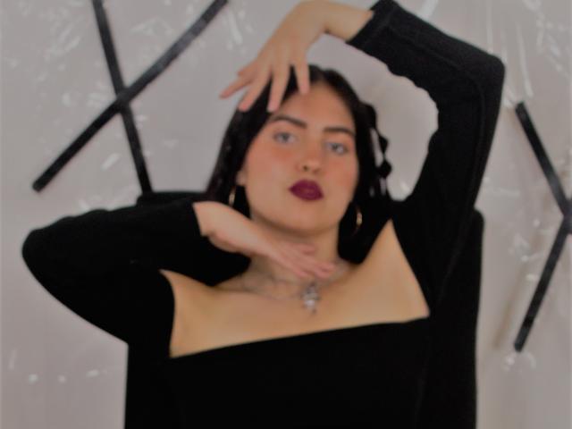 AgathaaQueen, con cabello oscuro, vistiendo una blusa negra de manga larga que deja ver sus senos, con los brazos levantados, una mano en la cabeza y otra en el cuello, usando lápiz labial púrpura y un collar..