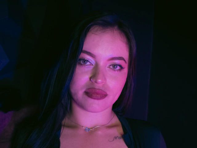 ZoeyStone with dark hair, nose ring, and purple lipstick, wearing a black top, has a small tattoo above her chest and a delicate necklace. Her eyes are slightly open.