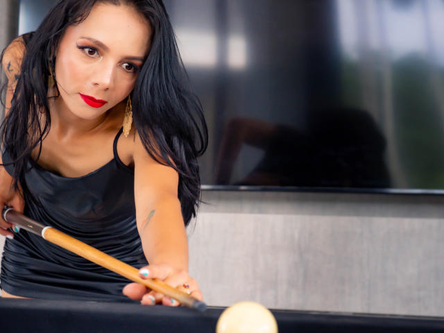 EvelinLuna with black hair and red lipstick, wearing a shiny black dress, leaning over a pool table, holding a cue stick, focused on a ball.
