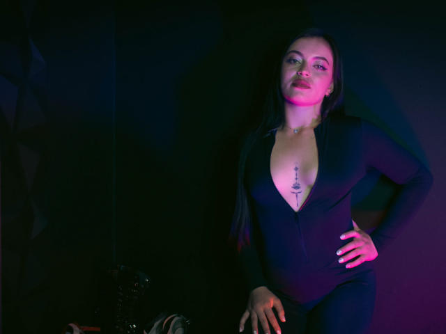 ZoeyStone, con lunghi capelli scuri, indossa una tuta nera con zip, che rivela un tatuaggio tra i seni, con le mani sui fianchi, illuminata da luci rosa e blu..