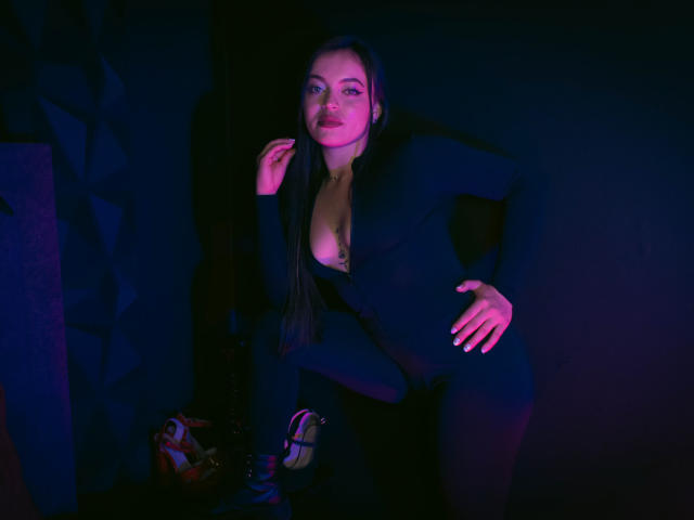 ZoeyStone with long dark hair, black outfit, and cleavage, kneeling, hand on hip, pink and blue lighting, showing tits, sexy, confident, hand near face, shoes in foreground.