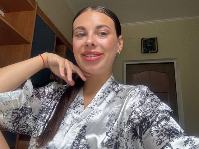 Mujer de raza negra, con senos medianos, vestida con una bata de seda floral plateada, cabello castaño recogido en una coleta, lápiz labial rojo, sonriendo, mano derecha en la mejilla, estante de madera y puerta en el fondo..