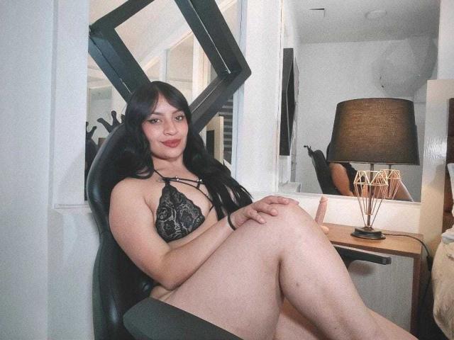 AlexaMiranda with long black hair, wearing a black lace bra, sitting on a chair, legs crossed, smiling at the camera, in a room with a lamp and mirror.